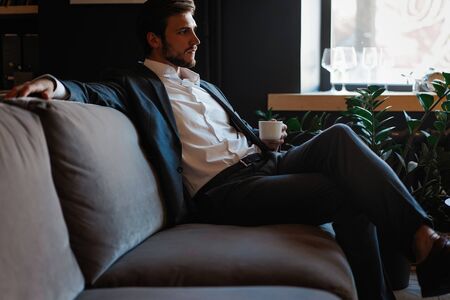 Handsome young man holding coffee cup sitting on the couch in office