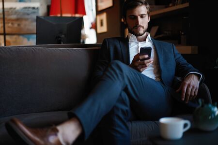 Smiling bearded man making text message on smartphone while sitting sofa at modern office.の写真素材