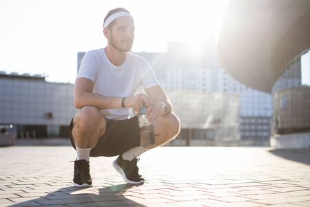 Young athlete in windrunner sitting on the street after good jogging session at sunset.