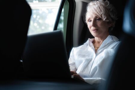 Senior businesswoman travelling to office in a car sitting on backseat with laptop.