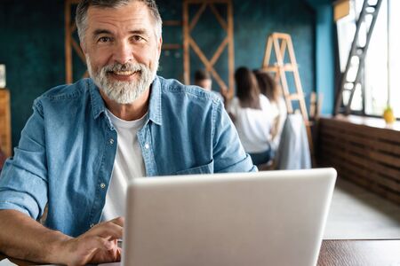 Cheerful mature man working on laptop and smiling while sitting at his working placeの写真素材