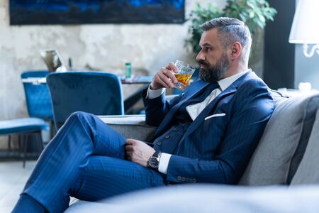 Handsome and successful businessman in stylish suit holding glass whiskey while sitting on the sofa at office.の写真素材