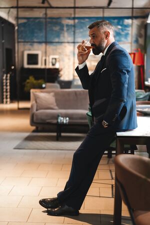 Mature handsome businessman stand and hold glass of whiskey in his own office.の写真素材