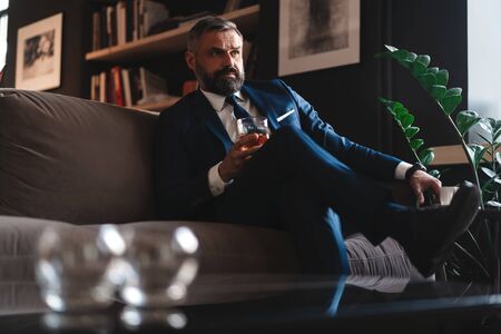 Handsome and successful businessman in stylish suit holding glass whiskey while sitting on the sofa at office.の写真素材
