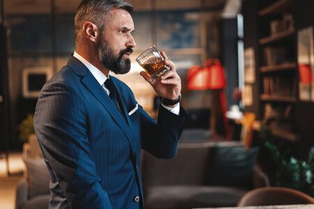 Mature handsome businessman stand and hold glass of whiskey in his own office.