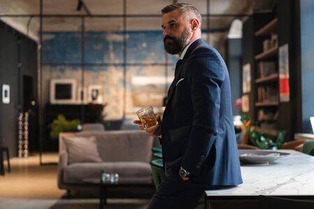 Mature handsome businessman stand and hold glass of whiskey in his own office.の写真素材