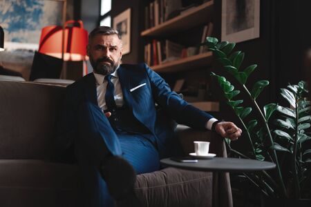 Starting new working day. Handsome man holding coffee cup sitting on the couch in office.の写真素材