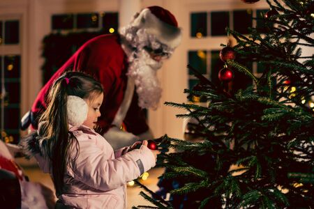 christmas, holidays and childhood concept - happy girl in santa hat decorating natural fir treeの写真素材