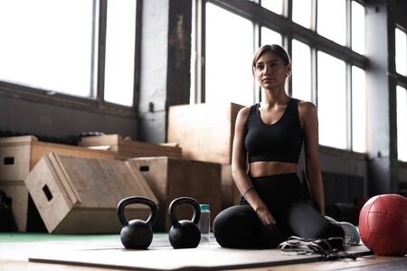 Young woman sitting on floor after her workout and looking down. Female athlete taking rest after fitness trainingの写真素材