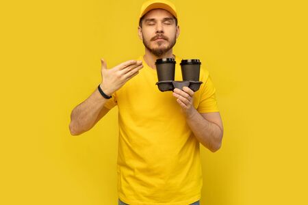 Portrait of cheerful delivery man in yellow uniform smiling and holding takeaway coffee cups...