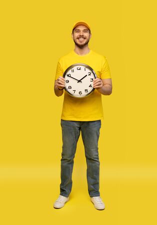 Delivery man in yellow workwear hold clock isolated on yellow background studio portrait.の写真素材