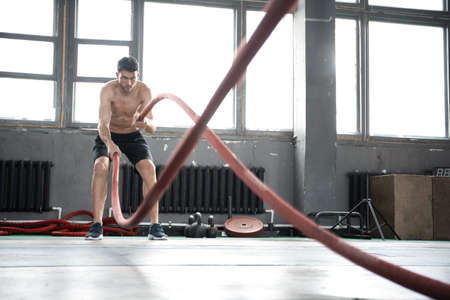 Athletic young man with battle rope doing exercise in functional training fitness gym.