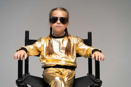 a little girl in gold suit sits on a chair. Isolated on a yellow background.の写真素材
