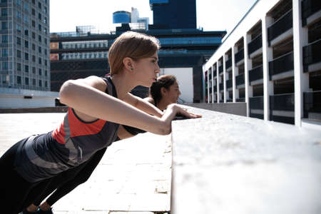Two attractive sport girls smiling while working out and doing push-ups in street city