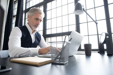 Concentrated at work. Mature man in full suit using laptop while working in modern office.