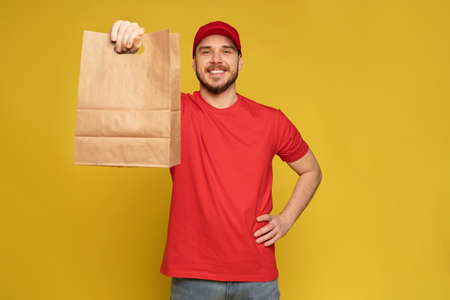 Man in red cap, t-shirt giving fast food order isolated on yellow background. Male employee courier...
