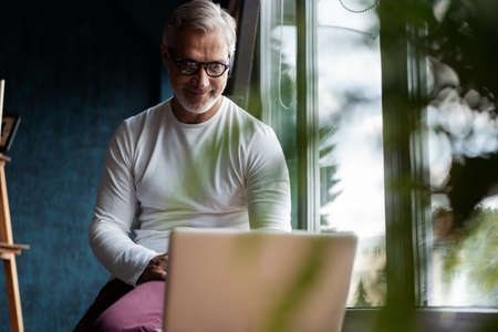 Casual Grey-haired Mature man in eyeglasses working on laptop while sitting in office or cafe near...