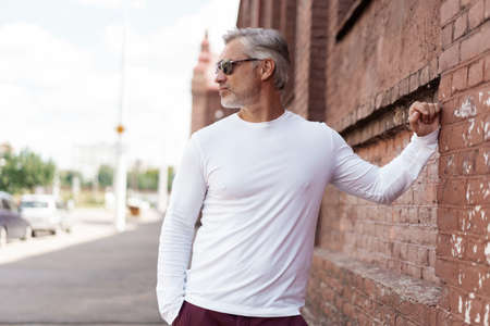 Portrait of a Grey-haired Mature handsome man in jeans and white t-shirt leaning to the old wallの写真素材