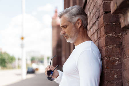 Portrait of a Grey-haired Mature handsome man in jeans and white t-shirt leaning to the old wallの写真素材