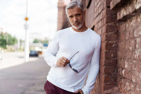 Portrait of a Grey-haired Mature handsome man in jeans and white t-shirt leaning to the old wall