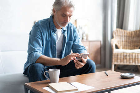 Senior man in couch reading message, using smartphoneの写真素材