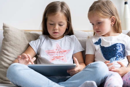 Little girls or sisrers sitting on the sofa playing digital tablet in the living room at home.の写真素材