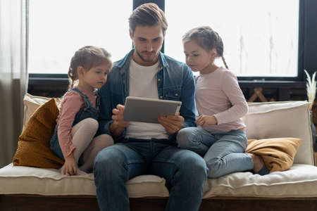 Beautiful young father, his cute little daughters are using a tablet and smiling, sitting on sofa at...