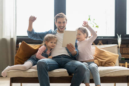 Beautiful young father, his cute little daughters are using a tablet and smiling, sitting on sofa at home.の写真素材