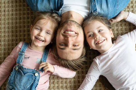 Top view of handsome young father and his cute little daughters looking at camera and smiling, lying on floor.の写真素材