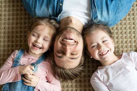 Top view of handsome young father and his cute little daughters looking at camera and smiling, lying on floor.の写真素材