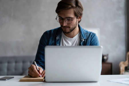 Serious young businessman with the glasses sitting and using laptop at home office and taking notes.