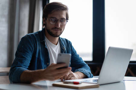 Young happy businessman smiling while reading his smartphone. Man working at his desk at home office