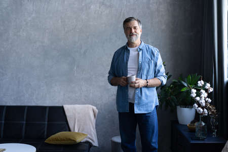 Handsome senior man is holding a cup, looking away and smiling while standing in apartment.