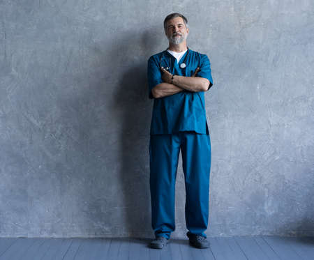 Full-length portrait of professional mature surgeon looking at camera while standing against the grey background.の写真素材