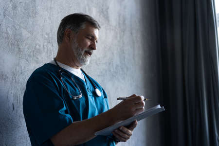Portrait of professional mature surgeon looking at camera while standing against the grey...