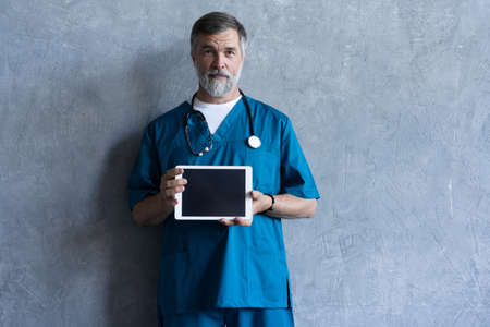 Portrait of professional mature surgeon holding a tablet, looking at camera while standing against the grey background.の写真素材