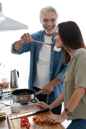 Beautiful couple. Man feeding wife and smiling while cooking in kitchen at home
