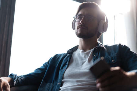 Smiling young adult man wearing wireless headphones listening mobile music on smartphone