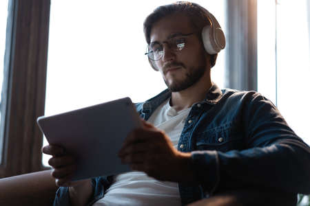 Handsome young man in casual clothes using a digital tablet and headphones and smiling while sitting on the chair.の写真素材
