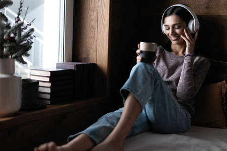 Beautiful young woman listening to music while sitting on sofa at home near the windowの写真素材