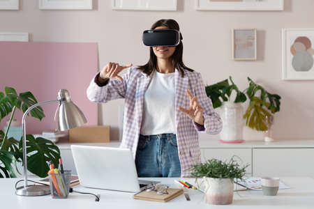 Confident young woman in virtual reality headset pointing in the air while standing at her working...