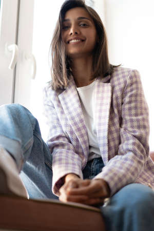 Young Indian mixed-race woman dressed casually sitting on the window sill at home officeの写真素材