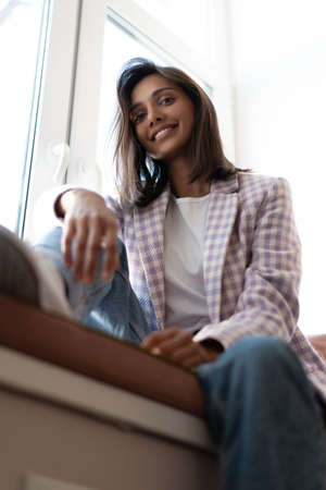 Young Indian mixed-race woman dressed casually sitting on the window sill at home officeの写真素材