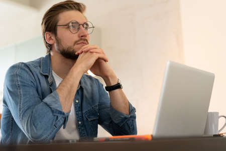 Young casual man working on laptop online, sitting at table in kitchen, looking at computer screenの写真素材