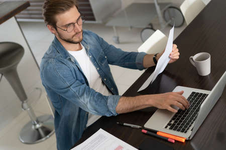 Young man working from home doing paperwork while using laptop.