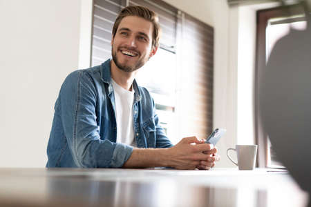 Confident man holding smartphone, sitting in kitchen, chatting online with friends or colleagues, browsing appの写真素材
