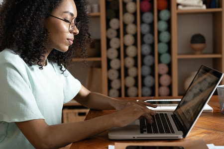 Young beautiful african american woman using her laptop while sitting in chair at her working placeの写真素材