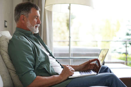 Portrait of happy mature man in casual clothes using laptop lying on sofa in house.
