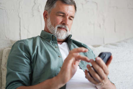 Smiling Senior Man Using Cellphone Browsing Internet Sitting On Sofa Indoor.