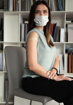 Portrait of a woman in protective mask receiving a vaccine, showing her arm with bandage after...